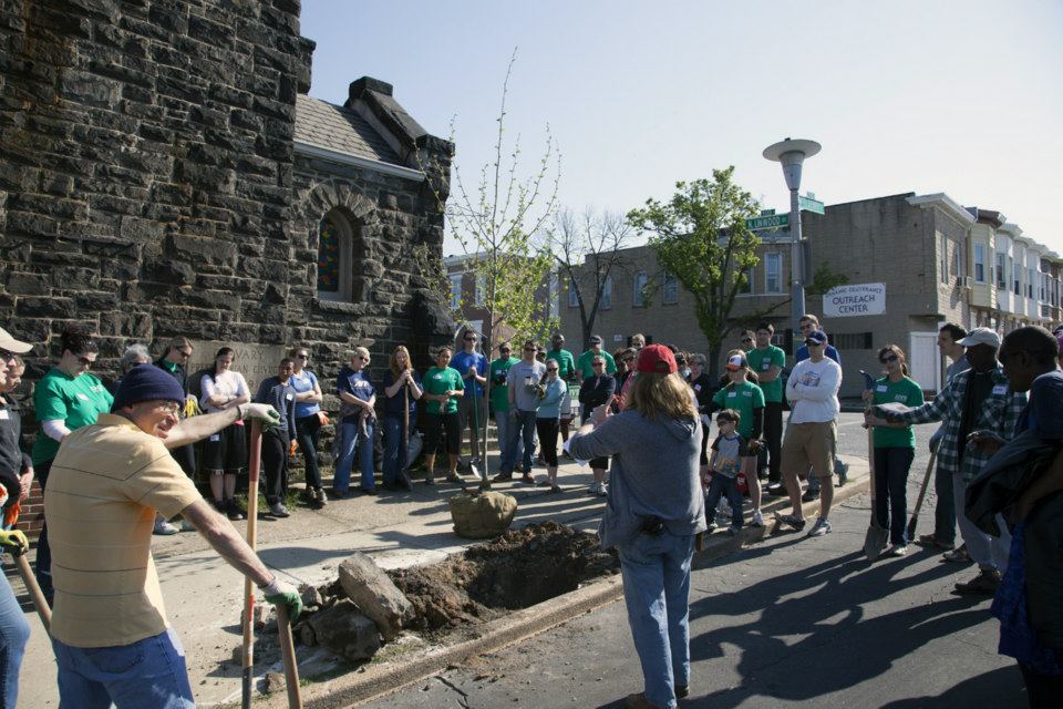 A volunteer and staff group gathering outside a stone building, with diverse people in casual clothing with shovels in hand. They are near a tree planting site.