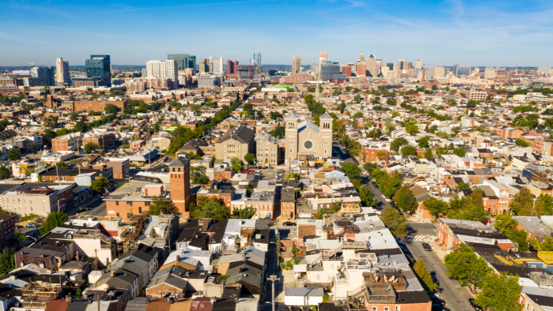 BaltimoreTreeTrust FAQ Aerial view of Baltimore's sprawling urban landscape, featuring tightly-packed residential buildings and a prominent church in the center. The city skyline is visible in the background under a clear blue sky, giving a vibrant and bustling feel.