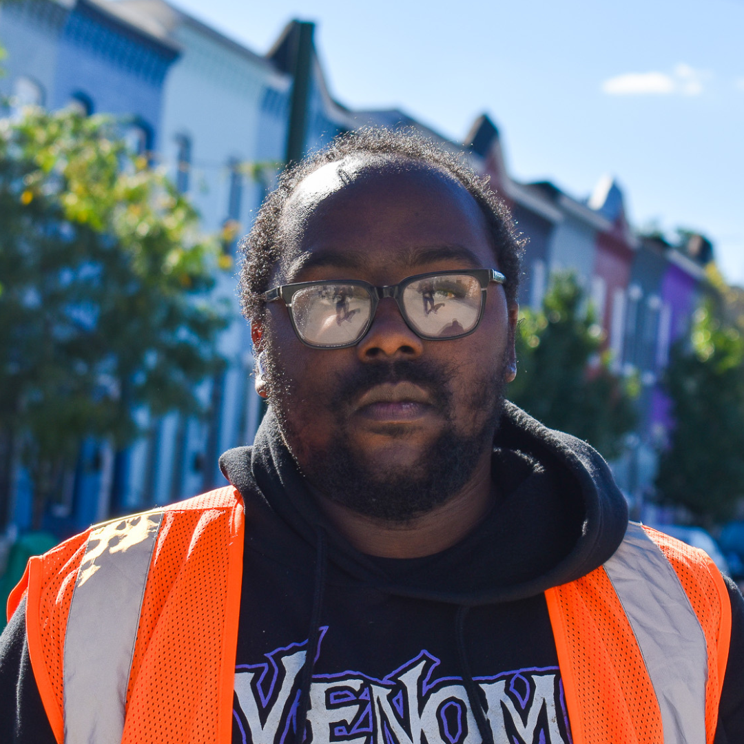 A bearded Black man wearing glasses, an orange safety vest, and a sweatshirt reading "Venom"