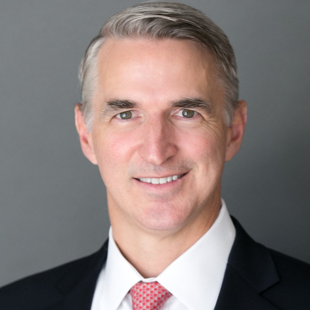 A smiling white man in a suit with a red tie stands against a gray background, conveying a professional and approachable demeanor.