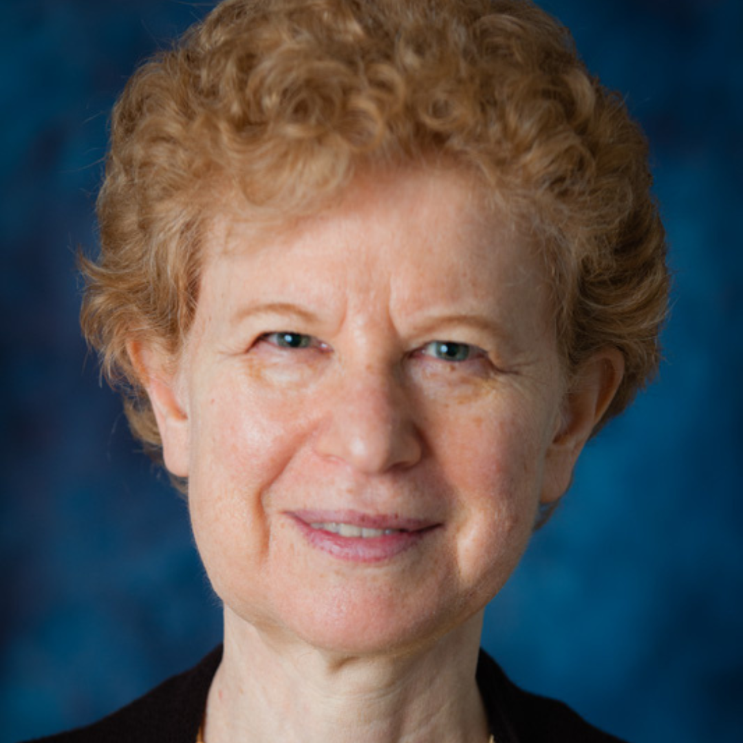 An older white woman with short, curly hair smiles slightly against a blue background. The image conveys a warm, approachable demeanor.
