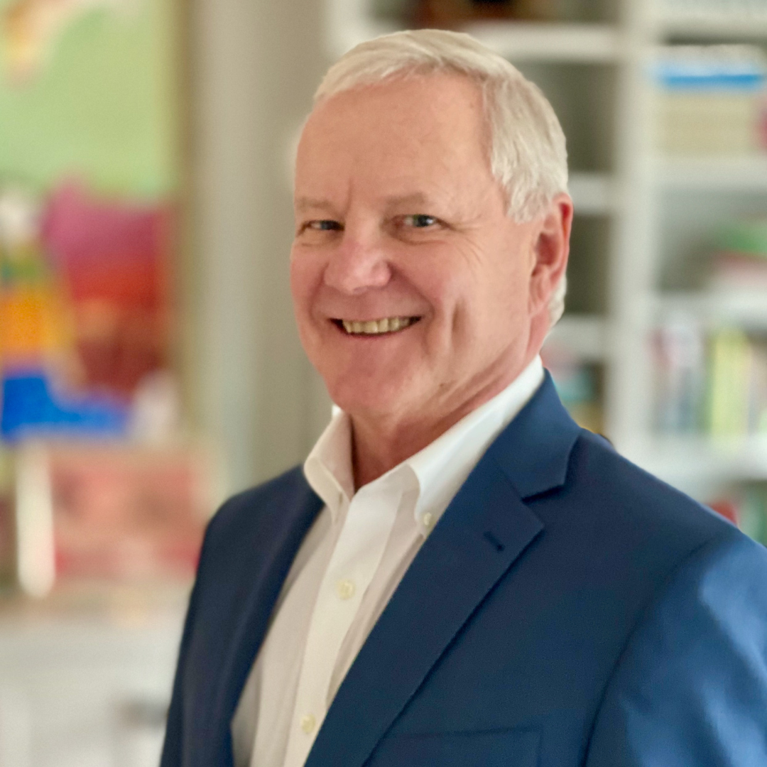 Smiling older white man in a blue suit and white shirt, standing in a well-lit room with colorful abstract art and bookshelves in the background.