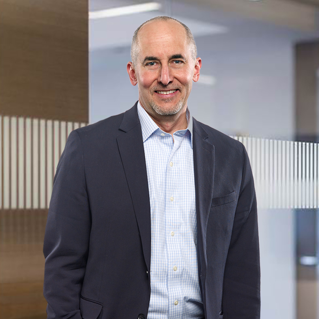 Smiling white man in a navy blazer and checkered shirt stands in a modern office. The background features glass panels and a warm, professional ambiance.